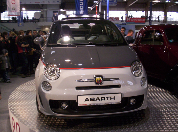 Ein roter und schwarzer Fiat 500 Abarth auf einer Autauschstellung mit Menschen drumherum und Bannern im Hintergrund.