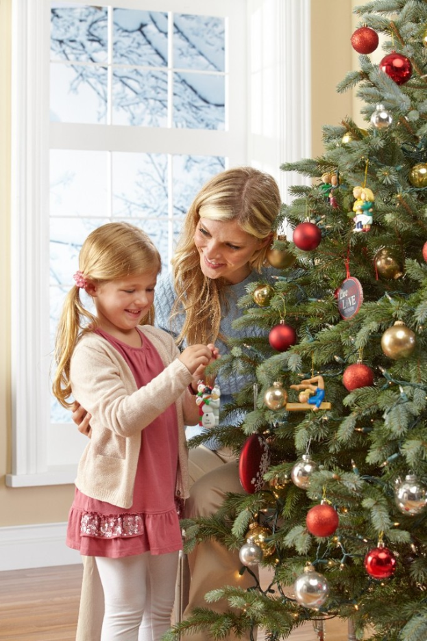 Ein geschmückter Weihnachtsbaum steht in einem Haus mit einer Frau und einem Kind im Vordergrund, einem Fenster und einer Wand im Hintergrund und Bäumen, dieoutside das Fenster sichtbar sind.