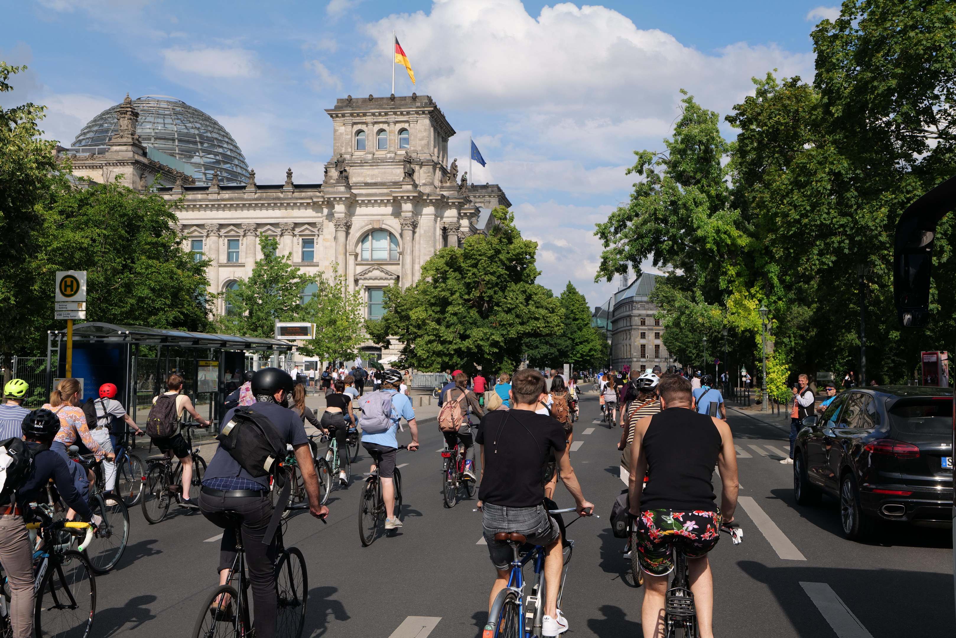 Eine Gruppe von Menschen, die Fahrräder auf einer von Bäumen gesäumten Straße in Berlin, Deutschland, fährt, mit einer Bushaltestelle auf der rechten Seite und einer Flagge, die auf einem nahegelegenen Gebäude weht.