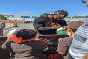 Cleveland Browns Wide Receiver Odell Beckham Jr. signiert Autogramme für Fans auf dem Trainingsgelände, umgeben von Menschen mit Mützen, einem Banner, Gebäuden, Bäumen und Zelten im Hintergrund bei einem bewölkten Himmel.