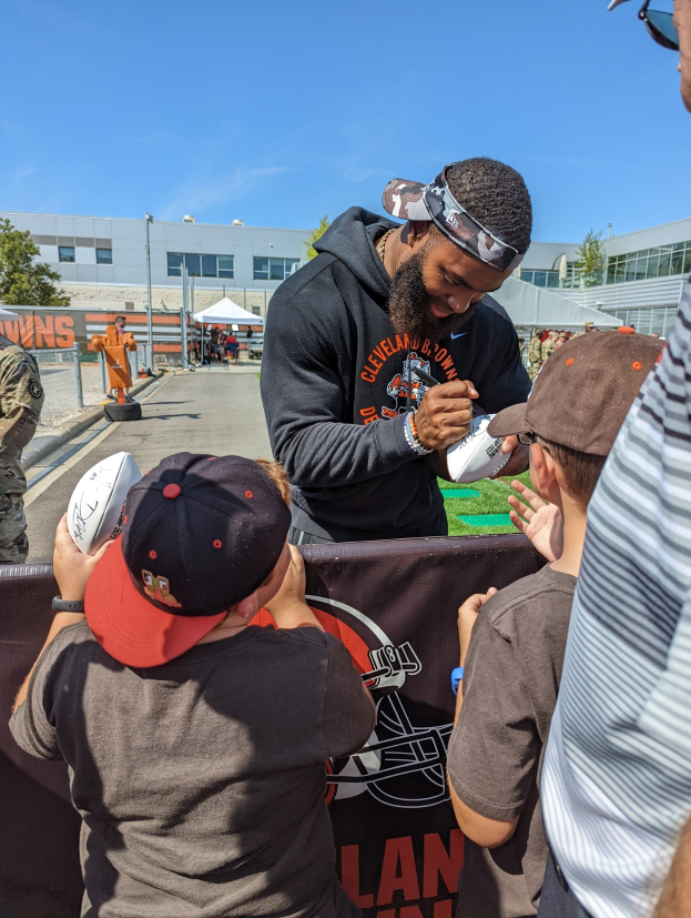 Cleveland Browns Wide Receiver Odell Beckham Jr. signiert Autogramme für Fans auf dem Trainingsgelände, umgeben von Menschen mit Mützen, einem Banner, Gebäuden, Bäumen und Zelten im Hintergrund bei einem bewölkten Himmel.