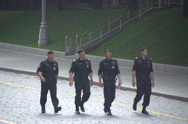 Vier Polizisten in Uniform gehen eine Straße entlang und halten Papiere in der Hand, mit einem Gebäude, Treppe, Geländer, Gras, Bäumen und einem Pfahl im Hintergrund.