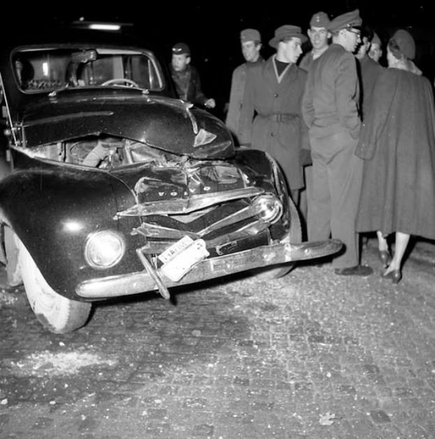 Ein Schwarz-Weiß-Bild eines auf der Straße liegenden Wagens mit einer Gruppe von Menschen drumherum, die das erhebliche Schadensbild am Kühlergrill und den zerbrochenen Fenstern begutachten.