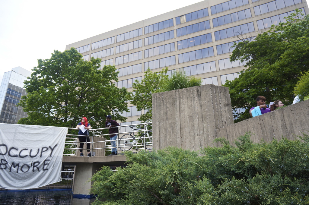 Menschen auf einer Brücke mit einem Banner und einem Fahrrad, unter der Wasser fließt, mit Bäumen, Gebäuden und Himmel im Hintergrund.