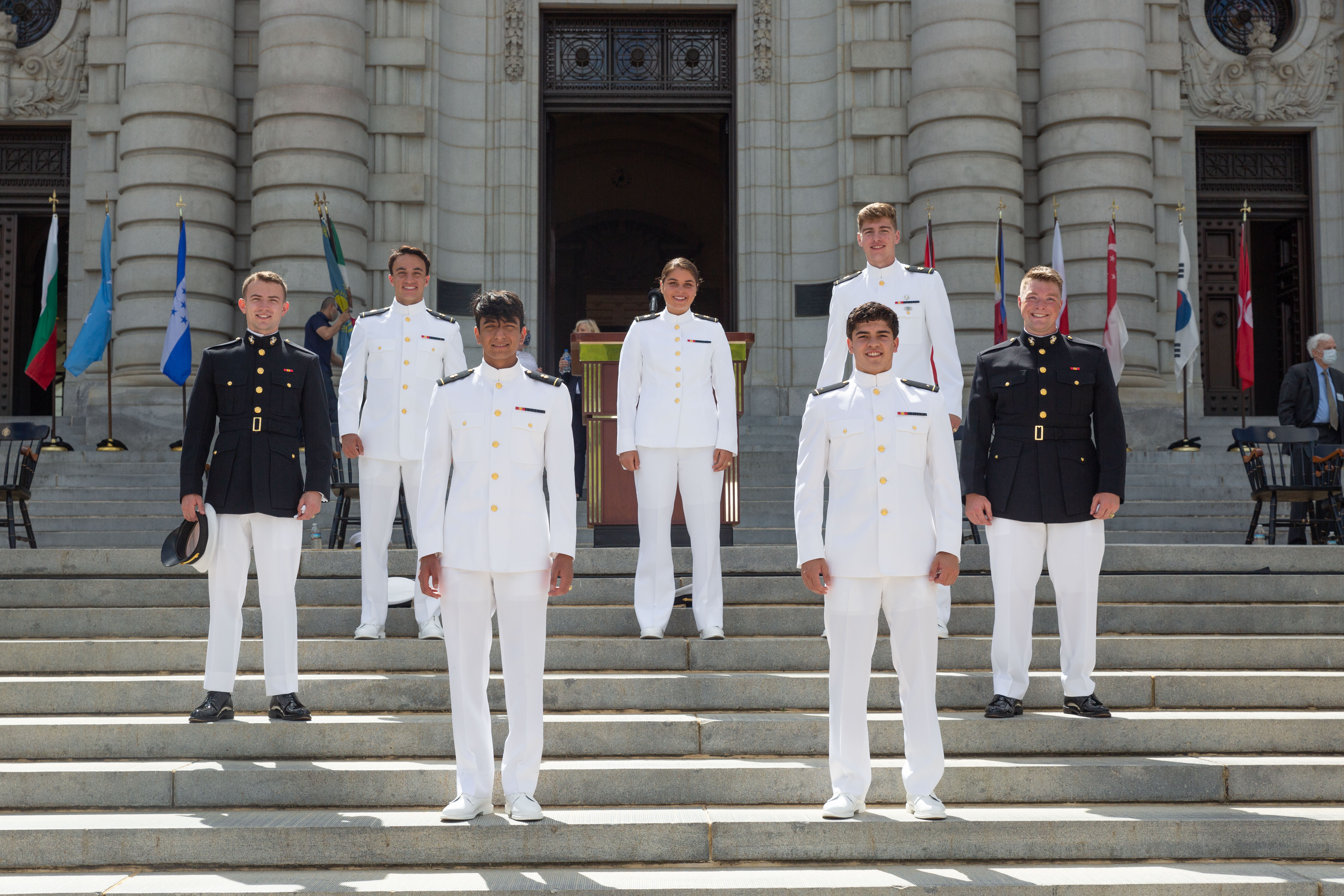 Eine Gruppe von Männern in weißen Uniformen auf einer Treppe, die lächeln und für ein Foto posieren, mit Menschen, Fahnen und Skulpturen im Hintergrund.