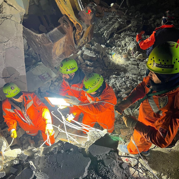 Eine Gruppe von Menschen in orangefarbenen Uniformen und grünen Helmen bei der Arbeit an Schutt mit einem Kran im Hintergrund, die Rettungsgeräte und Materialien halten.