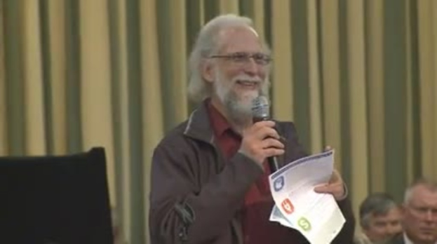 Eine Person steht an einem Podium und hält ein Mikrofon und Papier in der Hand, mit Stühlen und einem Vorhang im Hintergrund.