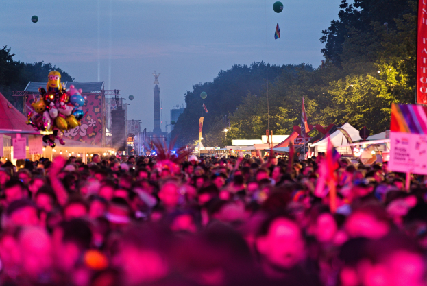 Eine große Menge auf einem nächtlichen Musikfestival mit bunten Lichtern, Ballons, Zelten, Bannern, Fahnen, Bäumen, einem Turm und einem sternenklaren Himmel im Hintergrund.