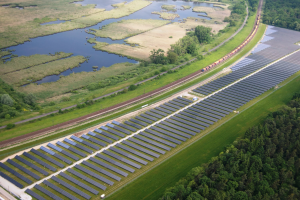 Luftaufnahme einer Solaranlage mit Panelen in einem Feld, umgeben von Bäumen, Gras, Wasser und einer nahen Bahntrasse.