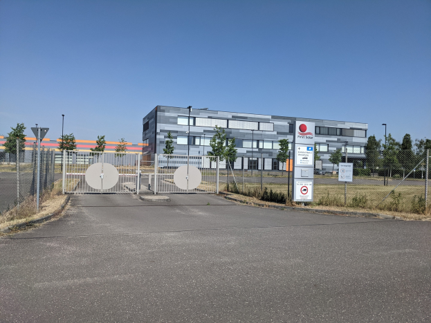 Großes industrielles Gebäude mit der Aufschrift "First Solar" und zahlreichen Fenstern, umgeben von einem Metallzaun, umgeben von Bäumen, Pflanzen, Gras, Schildern, Straßenmasten und einem Weg unter einem bewölktem Himmel.