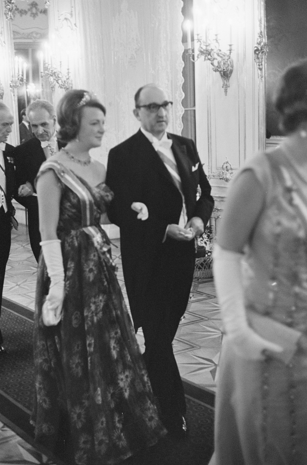 Ein Mann in einem schwarzen Blazer, weißem Hemd und schwarzen Hosen sowie eine Frau in einem eleganten Kleid gehen einen Flur entlang, umgeben von einer Gruppe von Menschen, die auf dem Boden stehen, mit ein paar an der Wand angebrachten Lampen, die eine warme Atmosphäre schaffen.