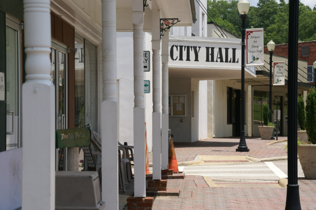 Rathaus mit umliegenden Gebäuden, Straßeninfrastruktur einschließlich Masten, Laternen und Namensschildern, Kraftfahrzeuge auf der Straße, Verkehrskegel, verstreute Stühle, Topfpflanzen, Bäume und eine bewölkte Kulisse.