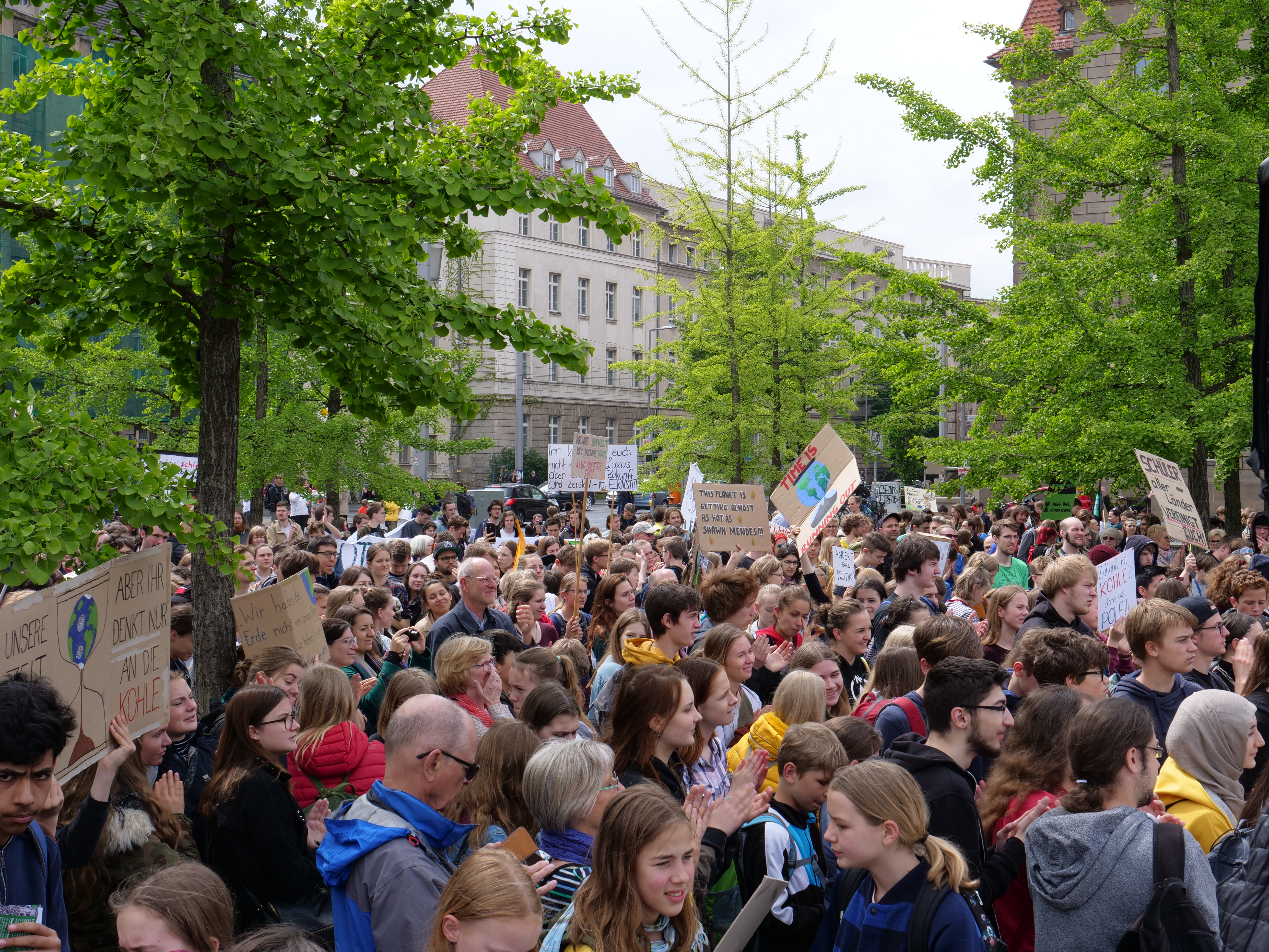 Eine große Menschenmenge mit Schildern steht vor einem Gebäude in Berlin, mit Bäumen, Fahrzeugen und einem Lautsprecher im Hintergrund.