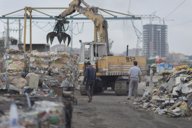 Eine Gruppe von Männern, die auf einem Schotterweg neben einem Haufen Müll gehen, mit einem Bagger im Hintergrund, Gebäuden, Bäumen, Polen, Drähten und anderen Gegenständen in der Umgebung.