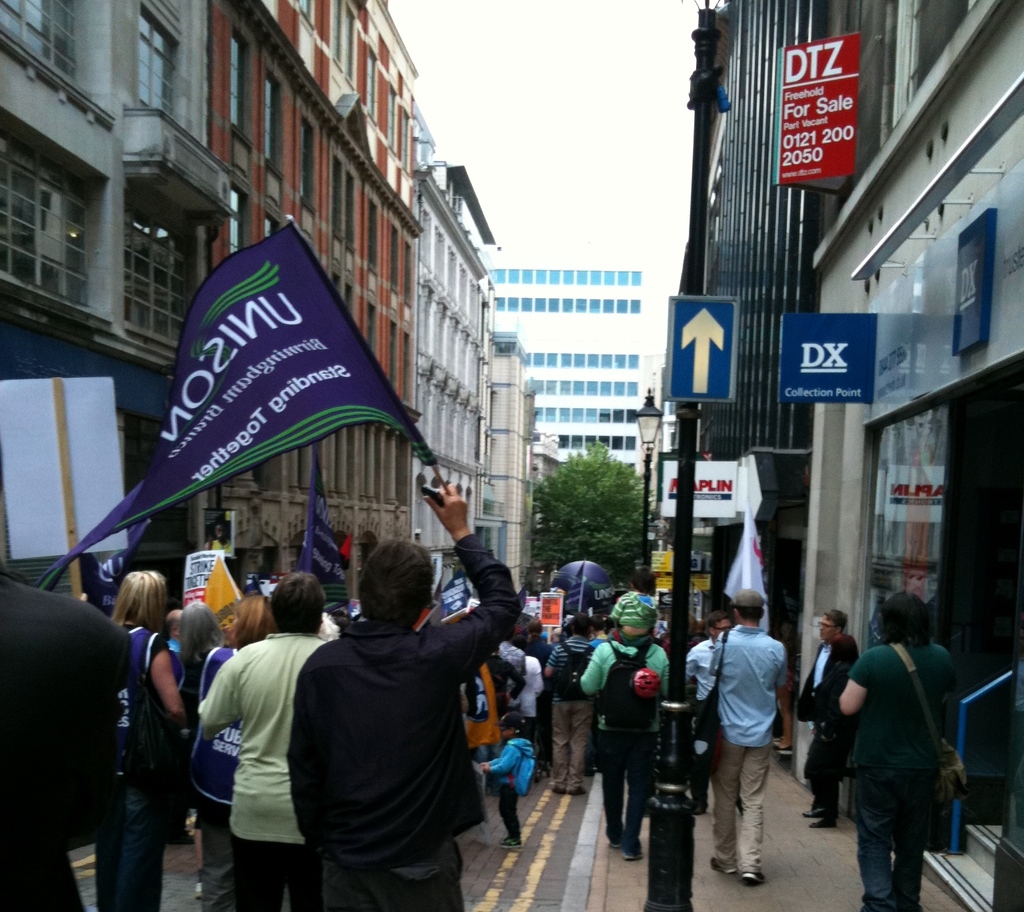 Eine große Gruppe von Menschen auf einer Straße, einige halten Fahnen und Schilder, mit einem Schildständer, Gebäuden mit Fenstern, einem Baum, einem Lichtmast und einem Gebäude im Hintergrund unter einem Himmel.