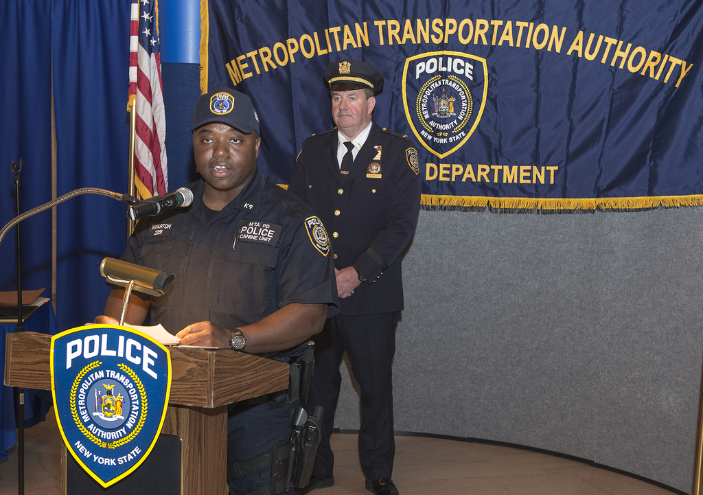 Zwei Polizeibeamte stehen vor einem Rednerpult, einer hält Papiere und spricht in ein Mikrofon, mit einer Tafel, Flagge und einem Tisch in der Nähe, und einer Tafel mit Symbolen und Text im Hintergrund.