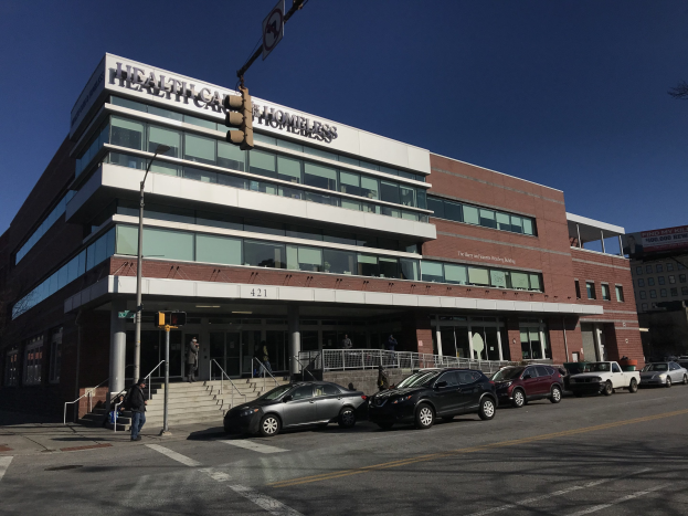 Großes Gebäude mit der Aufschrift "New York City Health Care Homeless Center" mit parkenden Autos, Fußgängern, Straßenmöblierung und Bäumen unter einem klaren Himmel.