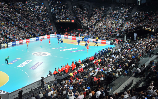 Eine Gruppe von Menschen, die Handball in einer großen Arena spielen, mit Zuschauern in den Rängen und Stufen, die zum Court führen.