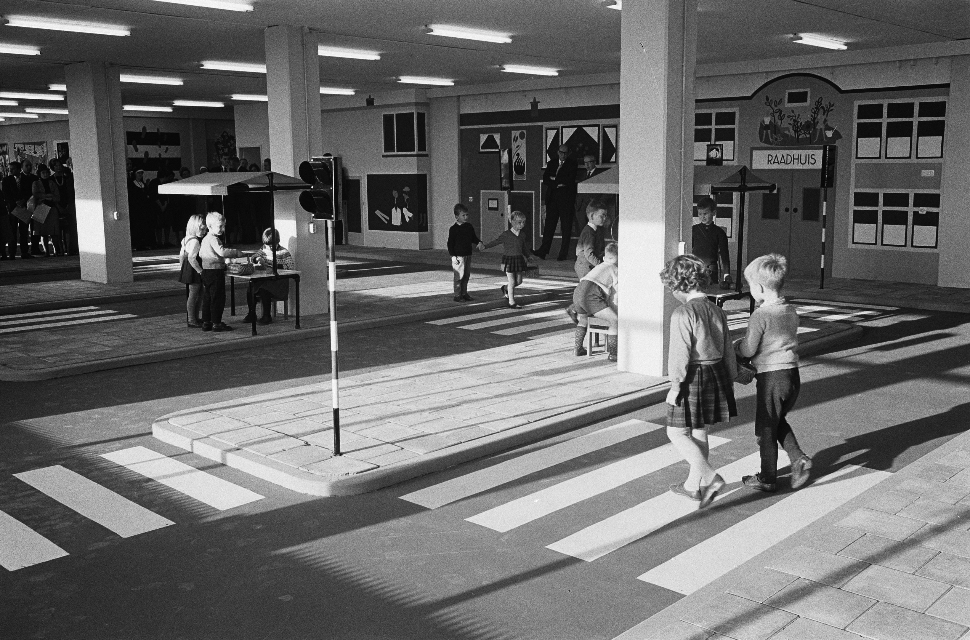 Schwarzes und weißes Foto von Kindern, die eine Straße an einer Fußgängerüberquerung überqueren, mit Fußgängern in der Nähe, einem Verkehrssignalmast im Vordergrund und strukturellen Säulen mit Deckenleuchten im Hintergrund.