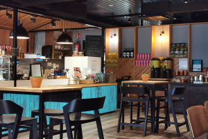 Innenansicht eines Restaurants mit Holztischen, Stühlen, einer Topfpflanze auf einem Tisch, einer Schiefertafel, einem Kühlschrank, einem Feuerlöscher, Regalen mit Flaschen, Schränken mit Gegenständen, einem gerahmten Foto an der Wand und Deckenleuchten.