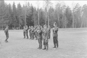 Schwarz-weißes Bild einer Gruppe von Männern in militärischer Kleidung, die in einem Feld stehen und Gewehre halten, mit Bäumen und Himmel im Hintergrund, wahrscheinlich während eines Trainingsübung.