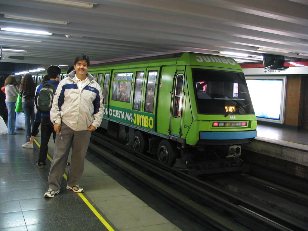 Ein Zug auf einem Bahngeleise mit Menschen, die links stehen und ein Dach oben im Bild.