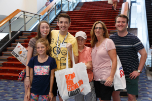 Eine Gruppe strahlender Menschen mit Taschen steht zusammen vor einer Rolltreppe, mit Treppen, Geländern und einer Wand im Hintergrund auf der Nationalen Buchmesse 2019.