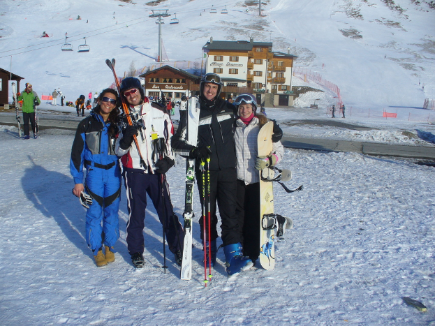 Vier Personen posieren mit Schlittschuhausrüstung und Werkzeugen vor einem Gebäude, anderen Menschen und einem schneebedeckten Berg.
