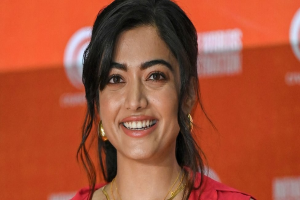 Eine Frau in einem roten Kleid mit einem eleganten Dutt und dezentem Make-up lächelt in die Kamera, trägt schwarze High Heels und hat ein gelbes Banner im Hintergrund mit der Aufschrift "Sonam Kapoors schönste Looks vom Cannes Film Festival".