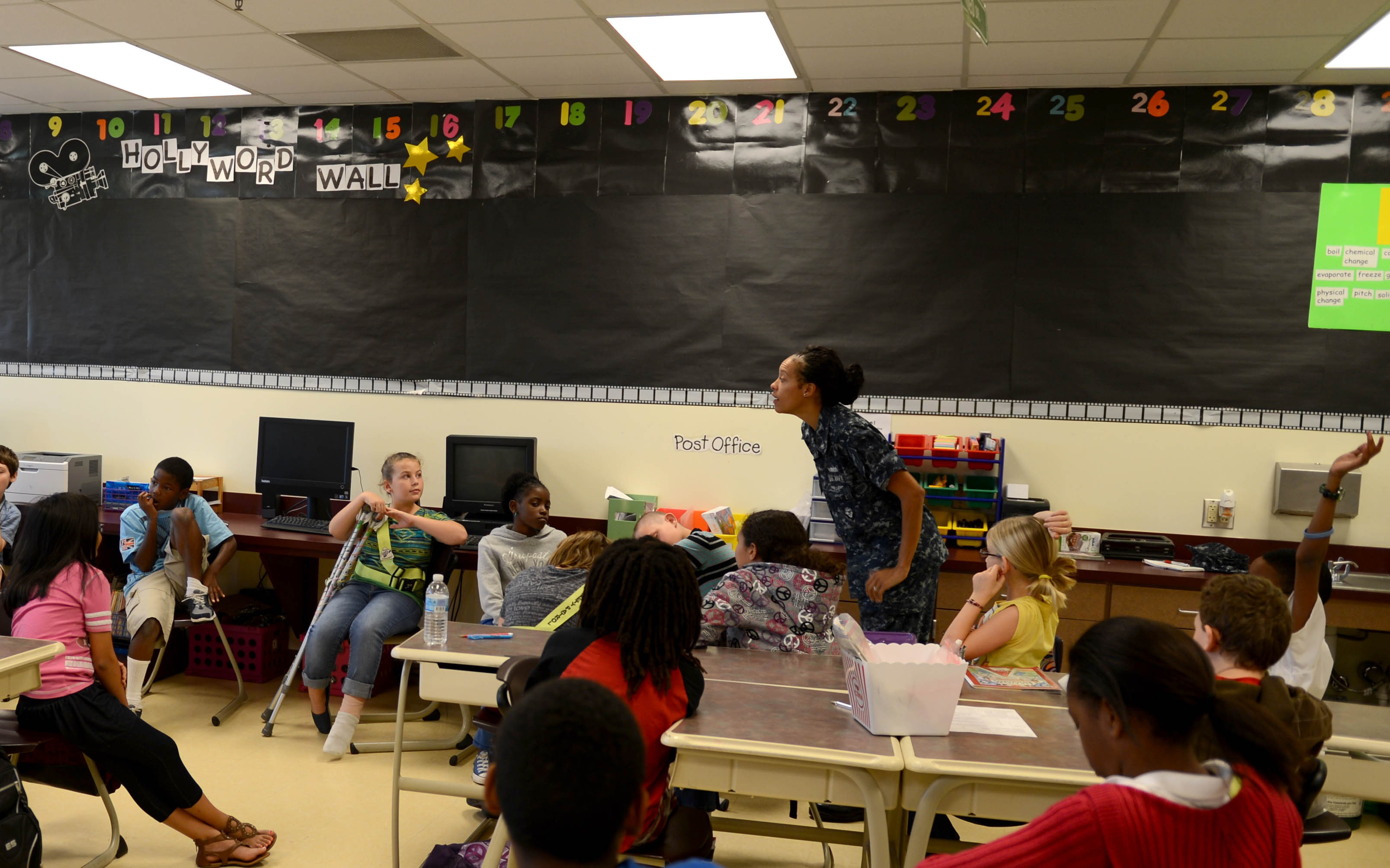 Eine Klasse mit Kindern an Tischen mit verschiedenen Gegenständen, einer Lehrerin in der Mitte und einer Tafel im Hintergrund.
