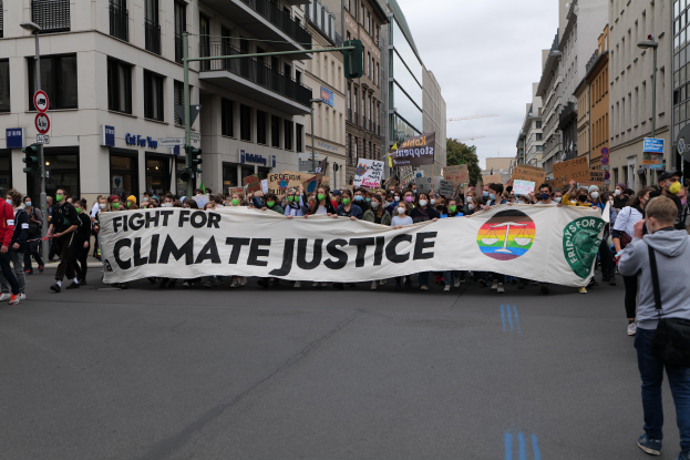 Eine Gruppe von Menschen marschiert mit einem "Kampf gegen die Klimagerechtigkeit"-Schild die Straße entlang, mit Gebäuden, Schildern, Ampeln und Bäumen an der Straße unter einem bewölkten Himmel.