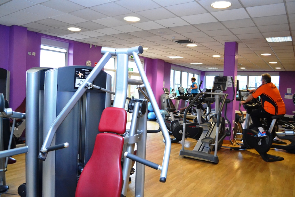 Ein Fitnessstudio mit mehreren Geräten und einigen Menschen beim Training, mit einer Decke und Lampen oben