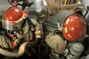 Feuerwehrleute in Schutzausrüstung arbeiten an einem Feuerhydranten mit Rohren und Ventilen im Hintergrund.