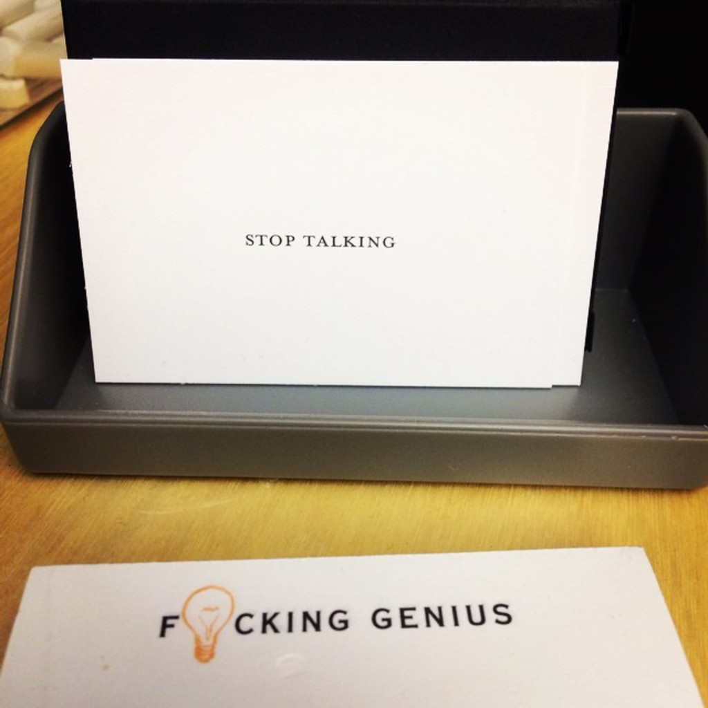 Ein Druckerbox mit einem Blatt Papier darauf, auf dem der Text 'Hör auf zu reden' steht, und einem weiteren Blatt auf dem Tisch, auf dem der Text 'Fucking genius' steht.