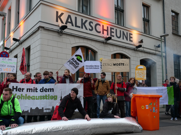Eine Gruppe von Menschen mit Schildern und Plakaten steht vor einem Gebäude, mit zwei Personen im Vordergrund und einem Müllcontainer auf der rechten Seite, während einer Protestaktion in Deutschland.