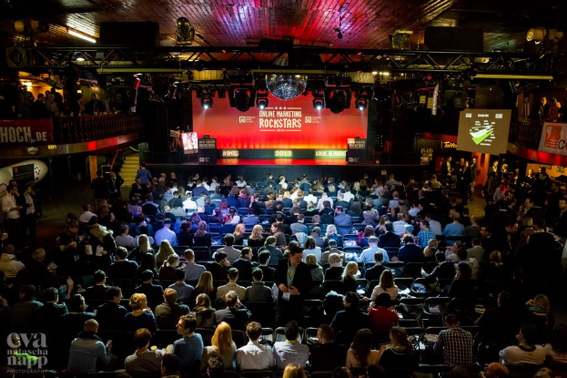 Eine große Menschenmenge sitzt auf Stühlen und schaut auf eine Bühne mit Bildschirmen und Plakaten, die von Scheinwerfern beleuchtet wird und von Geländern umgeben ist, mit einem Wasserzeichen in der linken unteren Ecke.