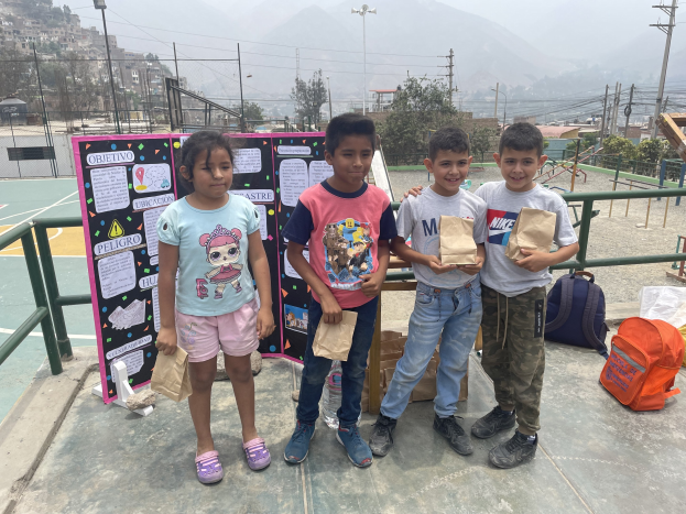 Gruppe von Kindern auf einem Basketballfeld, die Papiertüten halten, mit Tüten und Gegenständen rechts daneben und einer Tafel mit Text links daneben, vor einem Hintergrund aus Zaun, Pfosten, Straßenlaternen, Strommasten, Bäumen, Gebäuden, Hügeln und Himmel.