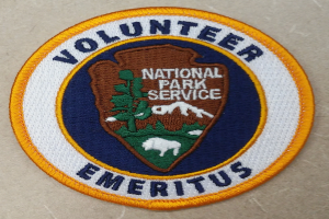 Ein rundes Patch mit einem blauen Rand, einem weißen Hintergrund und fettem schwarzem Text, der "Volunteer National Park Service Emeritus" in der Mitte eines gelben Kreises lesen.