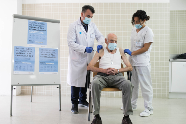 Ein älterer Mann sitzt in einem Stuhl und erhält von zwei maskierten und handschuhgetragenen Ärzten in einem Krankenhaus eine Impfung, wobei sich auf der linken Seite eine Tafel und auf der rechten Seite Gegenstände auf einer Plattform befinden.