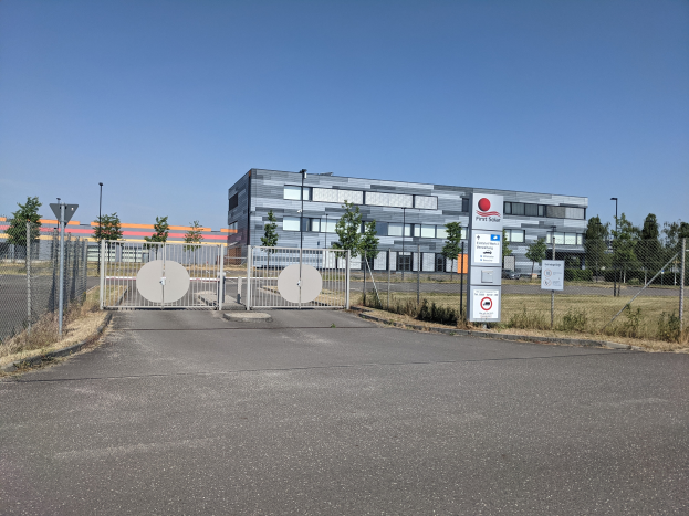 Ein großes Gebäude mit vielen Fenstern, beschriftet mit First Solar, umgeben von einem Metallzaun, Bäumen, Pflanzen, Gras, Schildern, Straßenmasten und einem Weg.