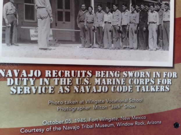 Ein Brett mit einer Gruppe von stehenden Menschen, einem Gebäude mit Fenstern, Pfählen und Text.