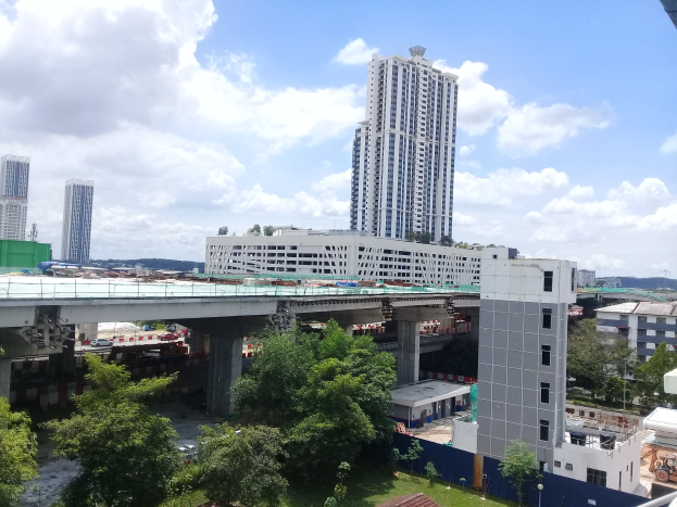 Luftaufnahme einer Stadtlandschaft mit Gebäuden, Bäumen, Gras, Fahrzeugen, einer Brücke, Masten und einem bewölktem Himmel, mit einer Baustelle für ein neues Wohnhochhaus im Vordergrund.
