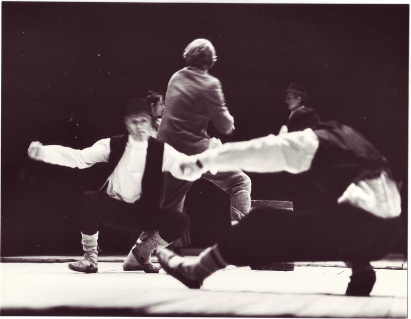 Eine Gruppe von Menschen, die auf einer Bühne tanzen, in einem Schwarz-Weiß-Foto, mit einer Person in der Mitte, die Schuhe trägt, vor einem dunklen Hintergrund.