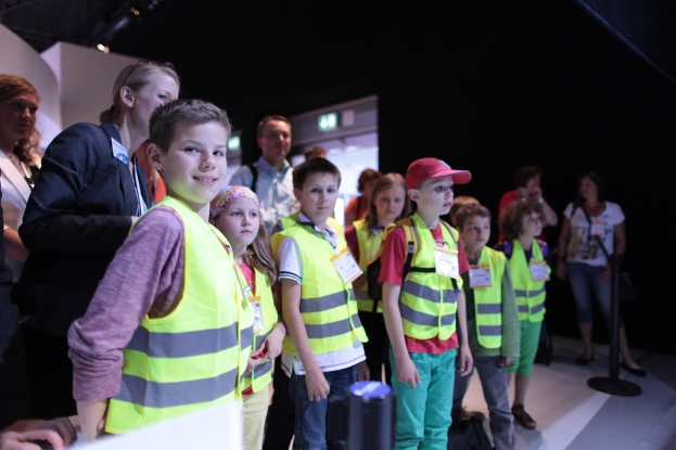 Eine Gruppe von Kindern mit Sicherheitswesten und Namensschildern, wobei ein Junge links lächelt, auf einem Boden mit einem Pfahl rechts und einigen Menschen im Hintergrund steht.