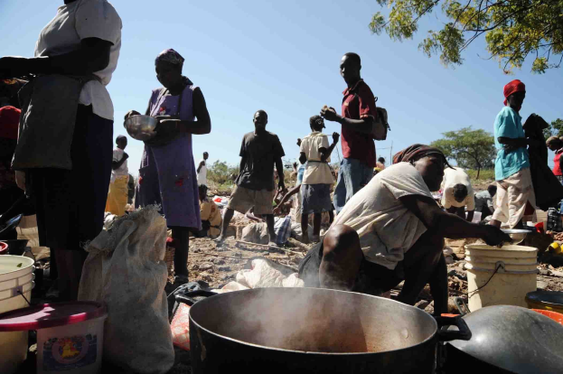 Eine Gruppe von Menschen, die Essen im Freien in einer ländlichen Gegend zubereiten, mit einem großen Topf, Eimern und Abdeckungen auf dem Boden, unter einem klaren blauen Himmel.