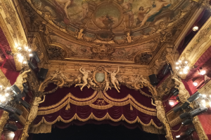Innere eines Theaters mit einem roten Vorhang, Kronleuchtern, Skulpturen, Lampen und einer Deckenmalerei, mit einer Bühne und ein paar Menschen unten.