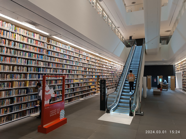 Große Bibliothek mit Bücherregalen in Reihen, eine Rolltreppe, eine Person, ein Textschild, sitzende Besucher auf Bänken, Säulen, Deckenleuchten und ein Wasserzeichen mit der Aufschrift "Shenzhen University Library."
