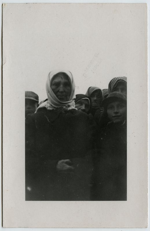 Ein Schwarz-Weiß-Foto einer Gruppe von Menschen, wahrscheinlich Flüchtlinge, die vor einer schmucklosen Wand stehen, wobei einige Individuals in traditioneller Kleidung und andere mit Hüten zu sehen sind.