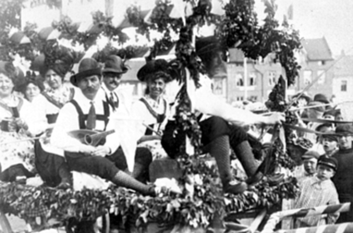 Schwarzes und weißes Bild eines Paradewagens mit Menschen in traditioneller bayrischer Tracht, einige spielen Instrumente, mit Blattdekoration, vorbei an Gebäuden unter einem klaren Himmel.