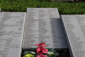 Denkmalstein mit Blumenstrauß darauf, umgeben von grünem Gras, mit Text, der wahrscheinlich Kriegsopfer gedenkt.